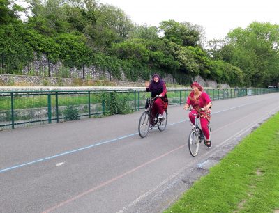 Two women ride bicycles in Preston Park. One of them waves.
